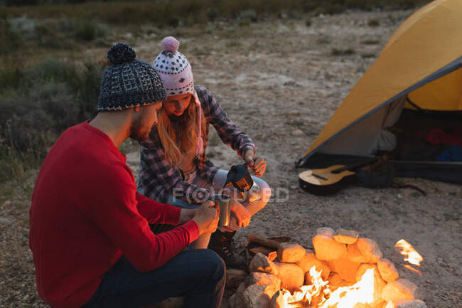 Seitenansicht eines kaukasischen Paares, das sich bei einem Ausflug in die Berge amüsiert, am Lagerfeuer sitzend, eine Frau gießt Kaffee in eine Metalltasse — Stockfoto