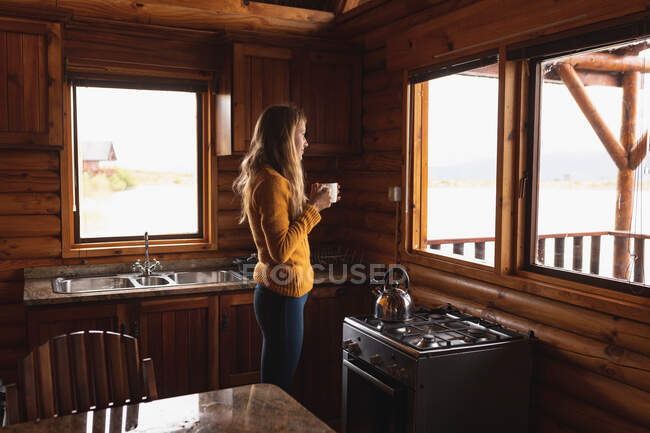 Vista frontal de una mujer caucásica pasándola bien en un viaje a las montañas, de pie en una cabaña de madera, bebiendo café, mirando por la ventana - foto de stock