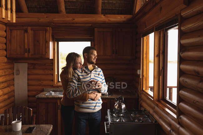 Vue de face d'un couple caucasien s'amusant lors d'un voyage à la montagne, debout dans une cabine, tenant une tasse de café, embrassant, regardant à travers la veuve — Photo de stock
