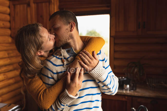 Vista frontal de una pareja caucásica pasándola bien en un viaje a las montañas, de pie en una cabaña, abrazando y besando - foto de stock