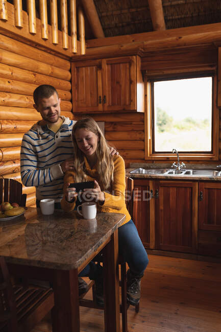 Front view of a Caucasian couple having a good time on a trip to the mountains, a woman is sitting by at table, showing something on a smartphone to a man standing behind her — Stock Photo