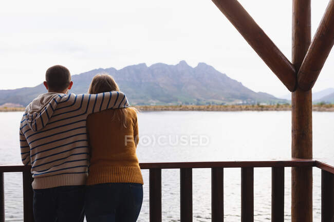 Rückansicht eines kaukasischen Paares, das sich auf einem Ausflug in die Berge amüsiert, auf einem Balkon in einer Hütte steht, sich umarmt, die Aussicht genießt — Stockfoto