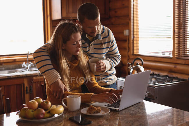 Seitenansicht eines kaukasischen Paares, das sich bei einem Ausflug in die Berge amüsiert, eine Frau sitzt und ihren Laptop benutzt, während ein Mann neben ihr steht und sie umarmt — Stockfoto