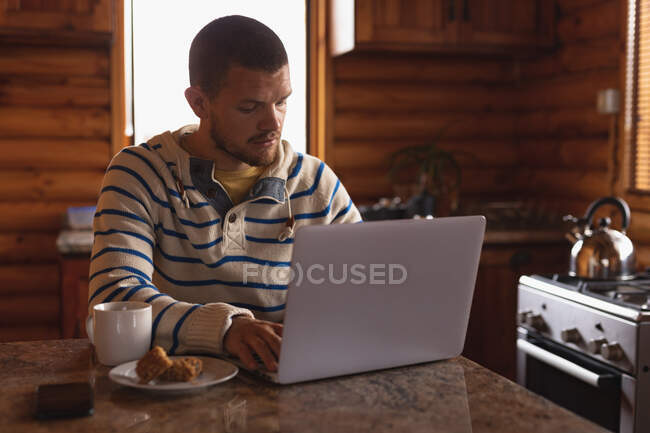 Передній вид на кавказького чоловіка добре проводить час у поїздці в гори, сидячи за столом у хатині, використовуючи ноутбук. — стокове фото