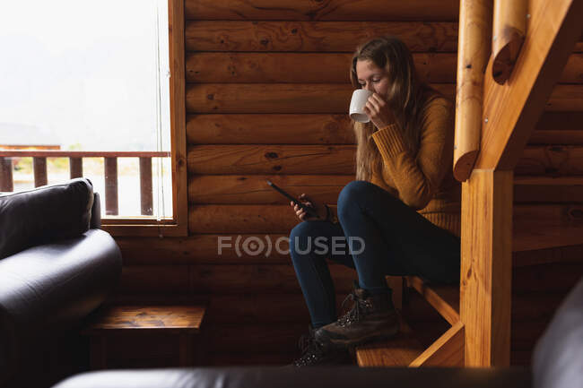 Vue de côté gros plan d'une femme caucasienne qui passe un bon moment en voyage à la montagne, assise sur les escaliers dans une cabine, utilisant un smartphone, buvant du café — Photo de stock