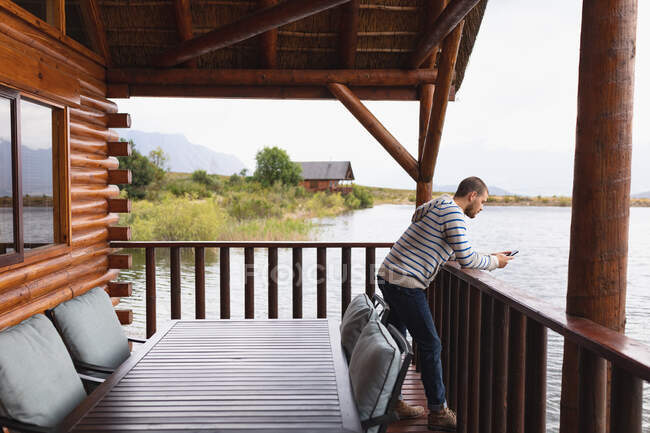 Вид сбоку на кавказца, хорошо проводящего время в поездке в горы, стоящего на балконе в каюте, опирающегося на барьер, используя смартфон — стоковое фото