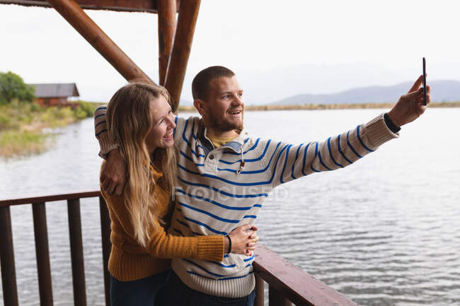 Vista laterale di una coppia caucasica che si diverte durante un viaggio in montagna, in piedi su un balcone in una cabina, abbracciando, prendendo un selfie — Foto stock