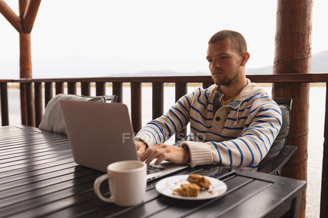 Vista lateral de um homem caucasiano se divertindo em uma viagem às montanhas, sentado a uma mesa em uma varanda em uma cabine, usando laptop — Fotografia de Stock