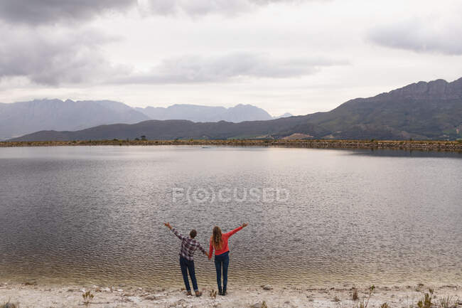 Передній вигляд кавказької пари добре проводить час у подорожі в гори, стоячи на березі озера, тримаючись за руки, піднімаючи руки вгору — стокове фото