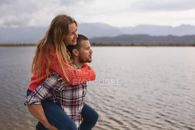 Vista lateral de cerca de una pareja caucásica pasar un buen rato en un viaje a las montañas, de pie en una orilla del lago, un hombre está llevando a una mujer cerdito espalda - foto de stock