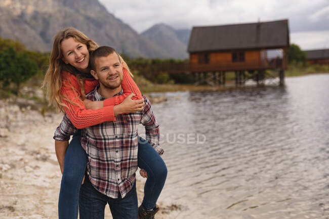 Vista frontal de cerca de una pareja caucásica pasando un buen rato en un viaje a las montañas, de pie en una orilla del lago, un hombre está llevando a una mujer cerdito - foto de stock