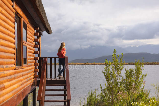 Vista laterale di una donna caucasica che fa un buon viaggio in montagna, in piedi su un balcone in una cabina, appoggiata su una barriera — Foto stock
