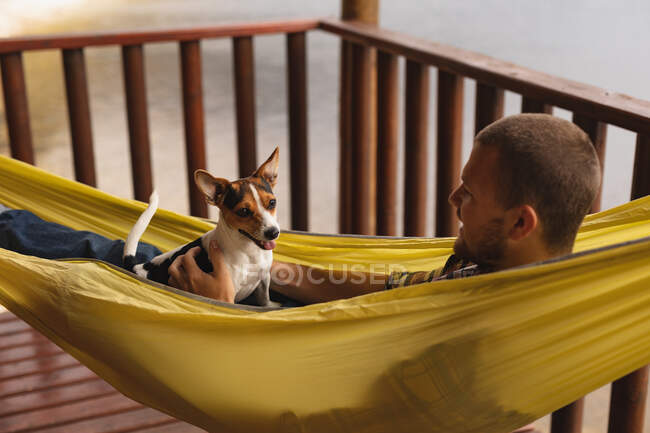 Vista lateral de perto de um homem caucasiano se divertindo em uma viagem às montanhas, deitado em uma rede com um filhote de cachorro em seu peito — Fotografia de Stock
