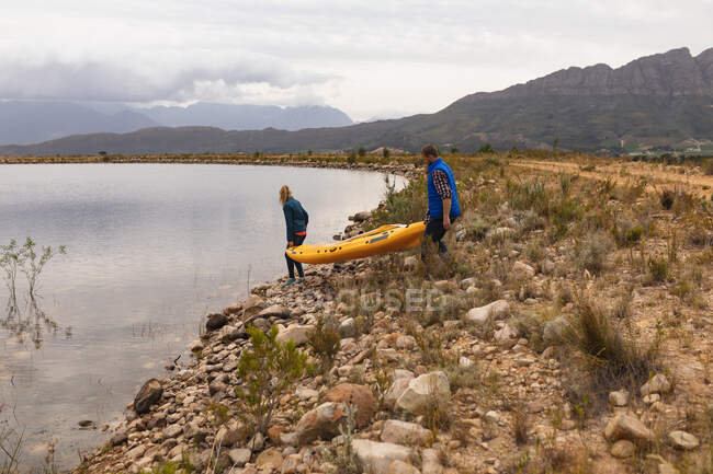 Vista lateral de una pareja caucásica pasándola bien en un viaje a las montañas, sosteniendo un kayak, preparándose para ponerlo en el agua - foto de stock