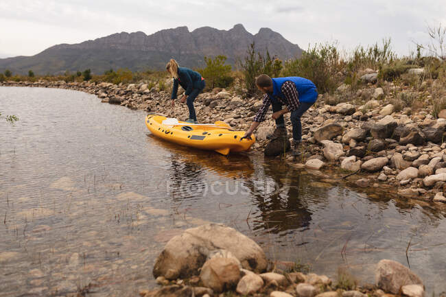 Vista lateral de um casal caucasiano se divertindo em uma viagem às montanhas, colocando um caiaque na água — Fotografia de Stock