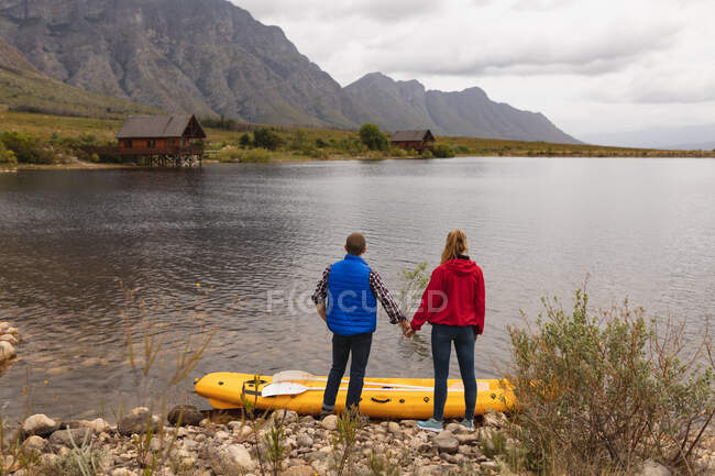 Side view of a Caucasian couple having a good time on a trip to the mountains, holding hands, enjoying their view after putting a kayak onto the water — Stock Photo