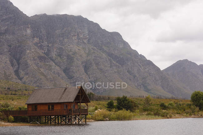 Breathtaking view of a cabin standing lonely by a lake with magnificent mountains behind it and a field between them — Stock Photo