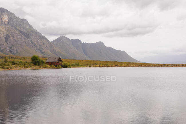 Breathtaking view of a cabin standing lonely by a lake with magnificent mountains behind it and a field between them — Stock Photo