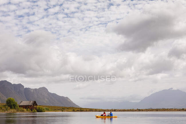 Vista lateral de una pareja caucásica pasando un buen rato en un viaje a las montañas, kayak en un lago, con una cabaña en el fondo - foto de stock