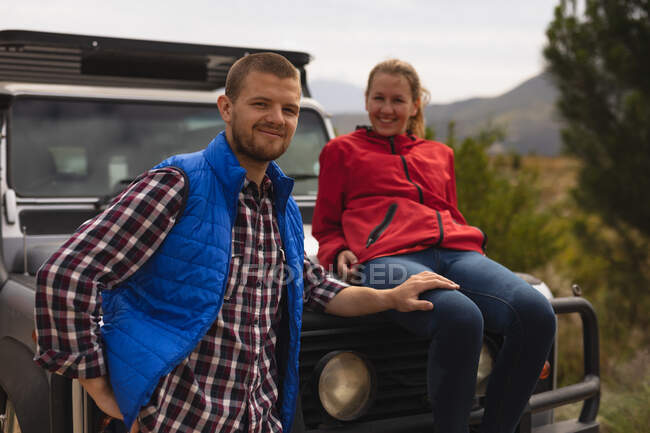 Vorderansicht eines kaukasischen Paares, das sich auf einer Reise in die Berge amüsiert, eine Frau sitzt auf einer Motorhaube und ein Mann hält ihren Oberschenkel und blickt in die Kamera. — Stockfoto
