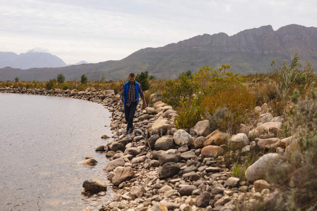 Vista frontal de un hombre caucásico pasándola bien en un viaje a las montañas, caminando sobre piedras en la orilla de un lago - foto de stock