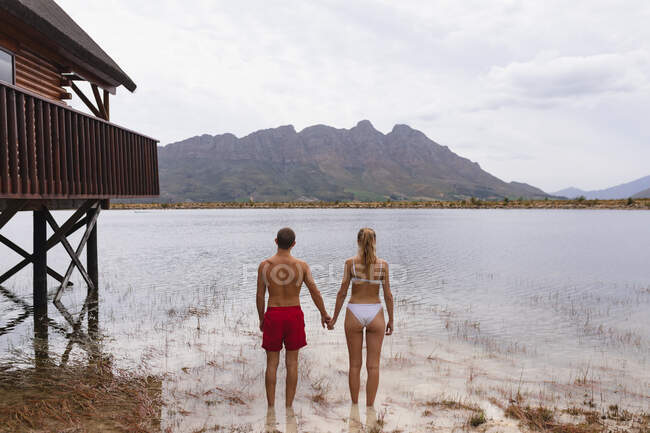 Vista trasera de una pareja caucásica pasándola bien en un viaje a las montañas, de pie en el lago, cogidas de la mano - foto de stock