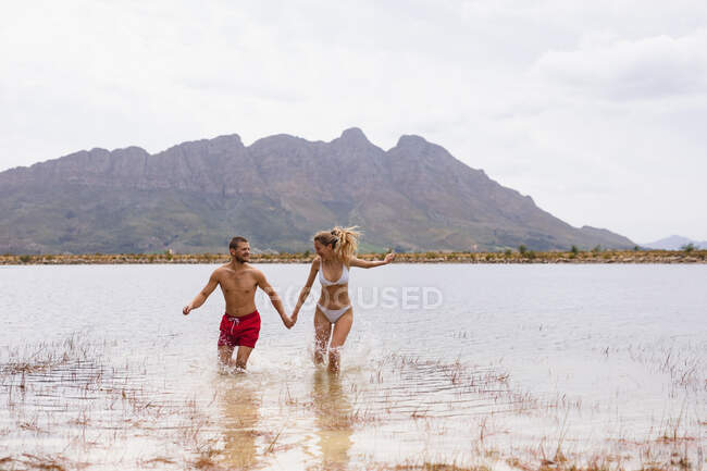 Vista frontal de una pareja caucásica pasándola bien en un viaje a las montañas, corriendo por el lago, tomados de la mano - foto de stock