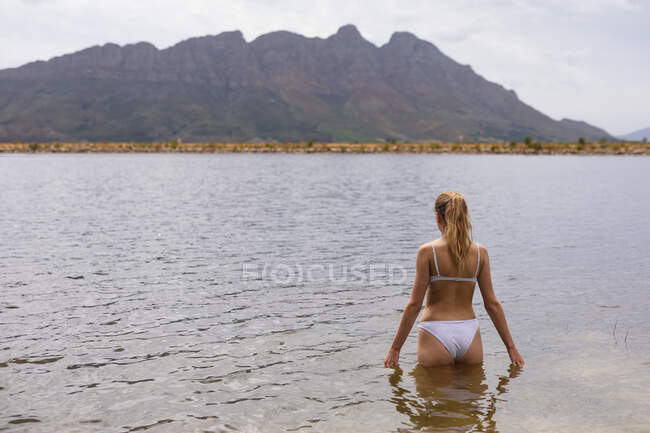 Rückansicht einer kaukasischen Frau, die sich bei einem Ausflug in die Berge amüsiert, in einem See steht und die Aussicht genießt — Stockfoto
