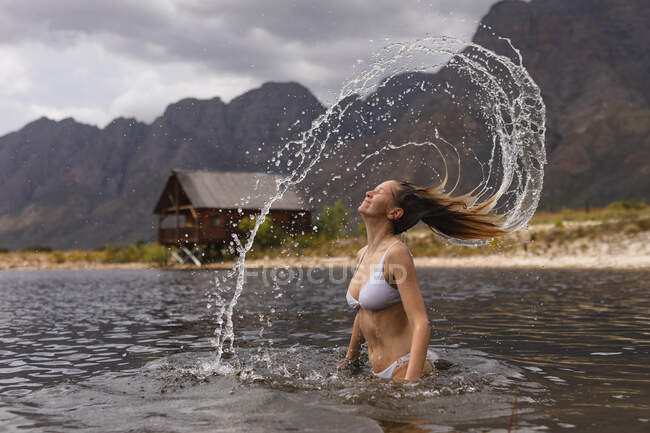 Rear view of a Caucasian woman having a good time on a trip to the mountains, standing in a lake, tossing her wet hair, leaving a water trail in the air — Stock Photo