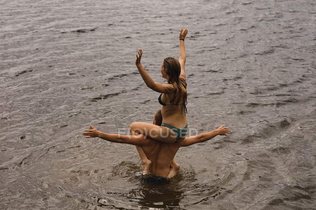 Vista trasera de ángulo alto de una pareja caucásica que se divierte en un viaje a las montañas, de pie en el lago, una mujer está sentada sobre los hombros de un hombre, con las manos en el aire - foto de stock
