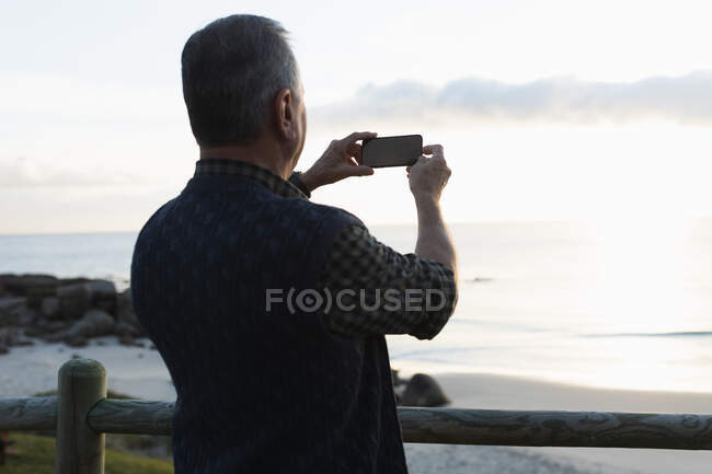Rear view of a senior Caucasian man standing alone by the sea by a fence, using his phone to take a photo — Stock Photo