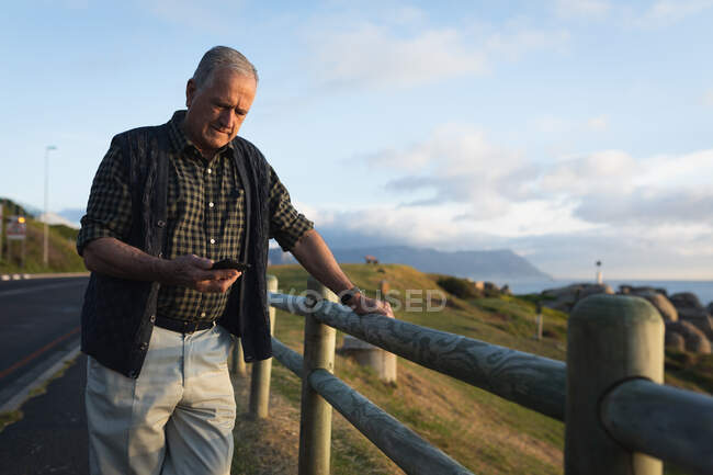 Vue de face d'un homme caucasien âgé debout seul au bord de la mer, utilisant son téléphone, penché sur une clôture au bord d'une route — Photo de stock