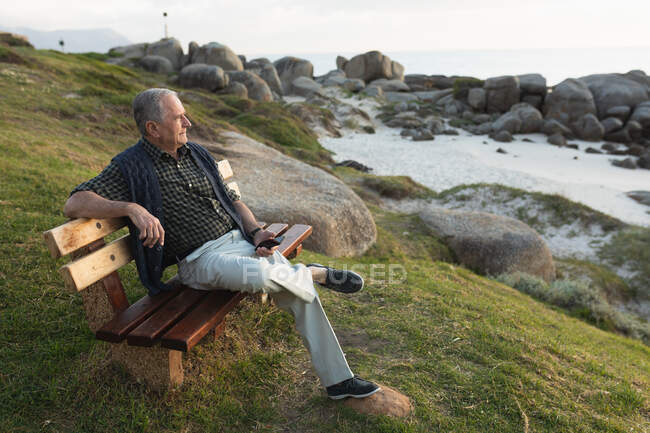 Vue latérale d'un homme caucasien âgé se relaxant, assis sur un banc au bord de la mer admirant une vue sur la côte — Photo de stock