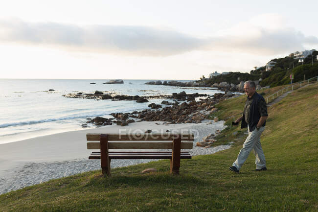 Vista laterale di un uomo caucasico anziano che cammina vicino al mare fino a una panchina, tenendo il suo smartphone — Foto stock