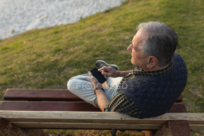 Vista de alto ângulo de um homem caucasiano sênior relaxante, sentado em um banco junto ao mar admirando uma vista costeira e usando um smartphone — Fotografia de Stock