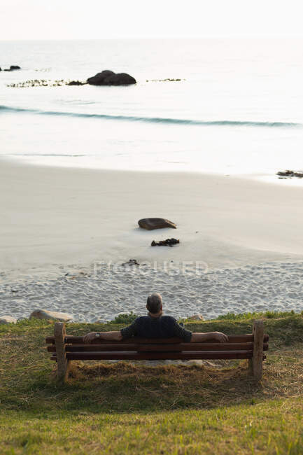 Задній вигляд високого кавказького чоловіка, який відпочиває, сидячи на лаві біля моря, милуючись краєвидом. — стокове фото