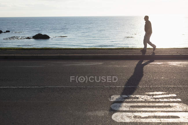 Вид сбоку на высокопоставленного кавказца, идущего в одиночестве по обочине дороги у моря, подсвеченной солнечным светом — стоковое фото
