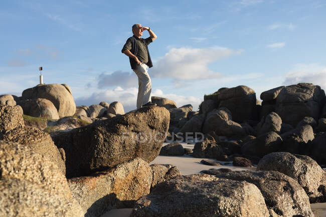 Seitenansicht eines älteren kaukasischen Mannes, der allein auf Felsen an einem Strand steht, seine Augen vor der Sonne abschirmt und einen Küstenblick bewundert, mit blauem Himmel im Hintergrund — Stockfoto