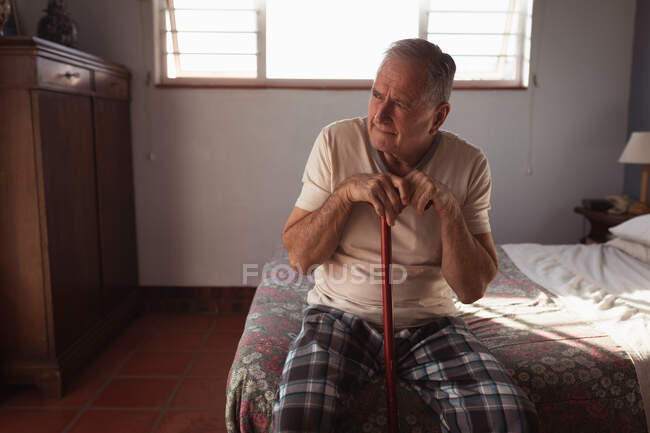 Передній погляд на старшого кавказького чоловіка відпочиває вдома в своїй спальні, сидячи на боці ліжка тримаючи тростину і думаючи після вставання вранці. — стокове фото