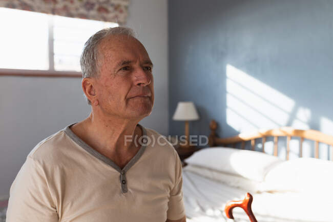 Vista lateral de cerca de un hombre caucásico mayor que se relaja en casa en su dormitorio, sentado en el lado de su cama con su bastón y pensando, después de levantarse por la mañana - foto de stock