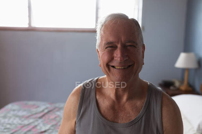 Ritratto di un anziano caucasico che si rilassa a casa nella sua camera da letto, indossa un gilet e guarda la telecamera sorridente, con una finestra illuminata dal sole dietro di lui — Foto stock