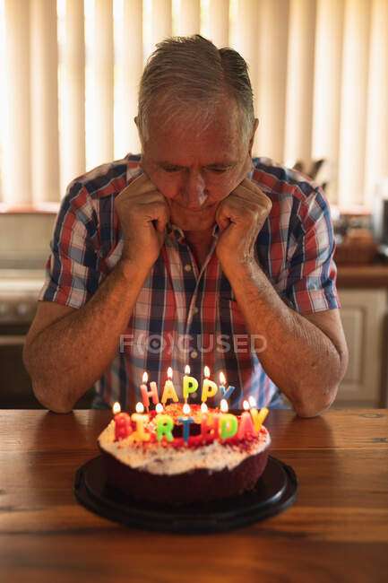 Вид спереди пожилого кавказца дома, сидящего в одиночестве за обеденным столом, смотрящего вниз на торт с зажженными свечами. — стоковое фото