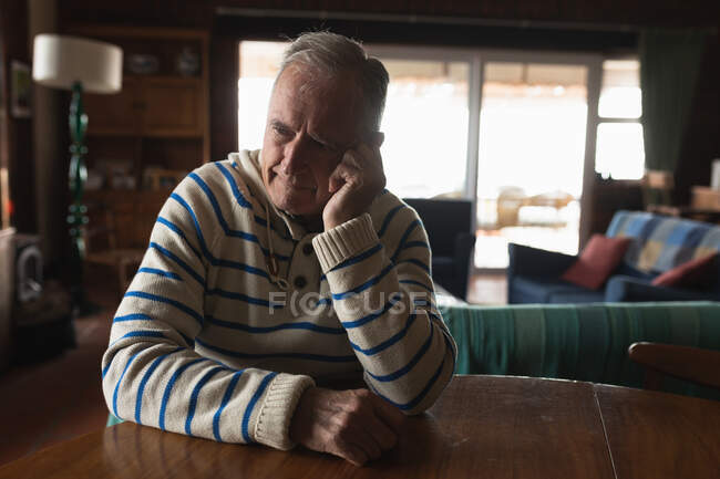 Vorderansicht eines älteren kaukasischen Mannes, der es sich zu Hause gemütlich macht, in seinem Esszimmer am Tisch sitzt, den Kopf in die Hand legt und nachdenkt — Stockfoto