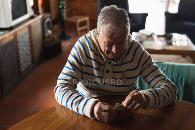 Вид спереди пожилого кавказца дома, сидящего за столом в столовой, смотрящего и трогающего деньги в бумажнике — стоковое фото