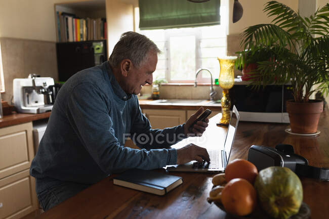 Vista lateral de un hombre caucásico mayor que se relaja en casa, sentado en el mostrador en su cocina usando un teléfono inteligente y computadora portátil - foto de stock