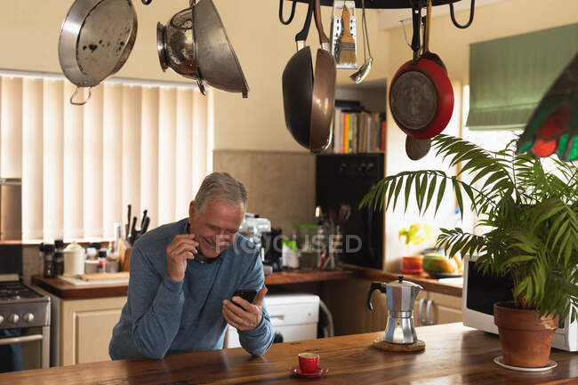 Vista frontale di un anziano caucasico che si rilassa a casa, seduto al bancone della sua cucina con uno smartphone e sorridente — Foto stock
