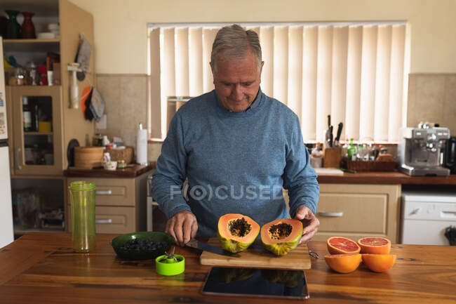 Вид пожилого кавказца, расслабляющегося дома, стоящего у прилавка на кухне, аккуратно разрезающего фрукты пополам острым ножом — стоковое фото
