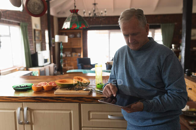 Vista frontal de un hombre caucásico mayor que se relaja en casa, sentado en el mostrador de su cocina, usando una tableta - foto de stock