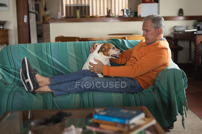 Side view of a senior Caucasian man relaxing at home in his living room, sitting on the sofa with his legs up playing with his pet dog — Stock Photo