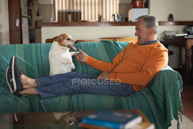 Vista laterale di un uomo caucasico anziano che si rilassa a casa nel suo soggiorno, seduto sul divano con le gambe alzate a giocare con il suo cane da compagnia — Foto stock